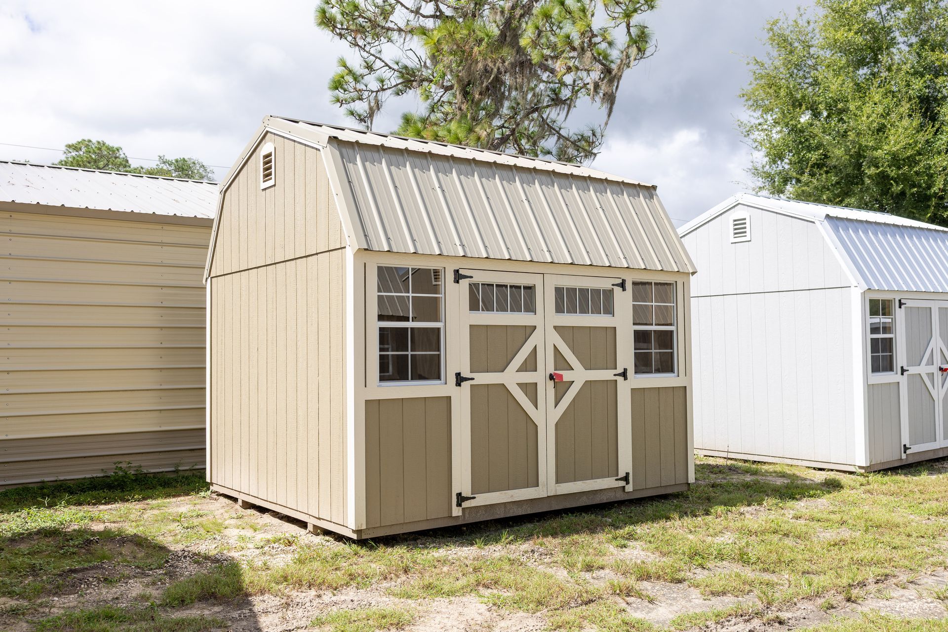 Tan shed with barn-style roof and double doors; two windows. Other sheds in background. Outdoors, sunny.
