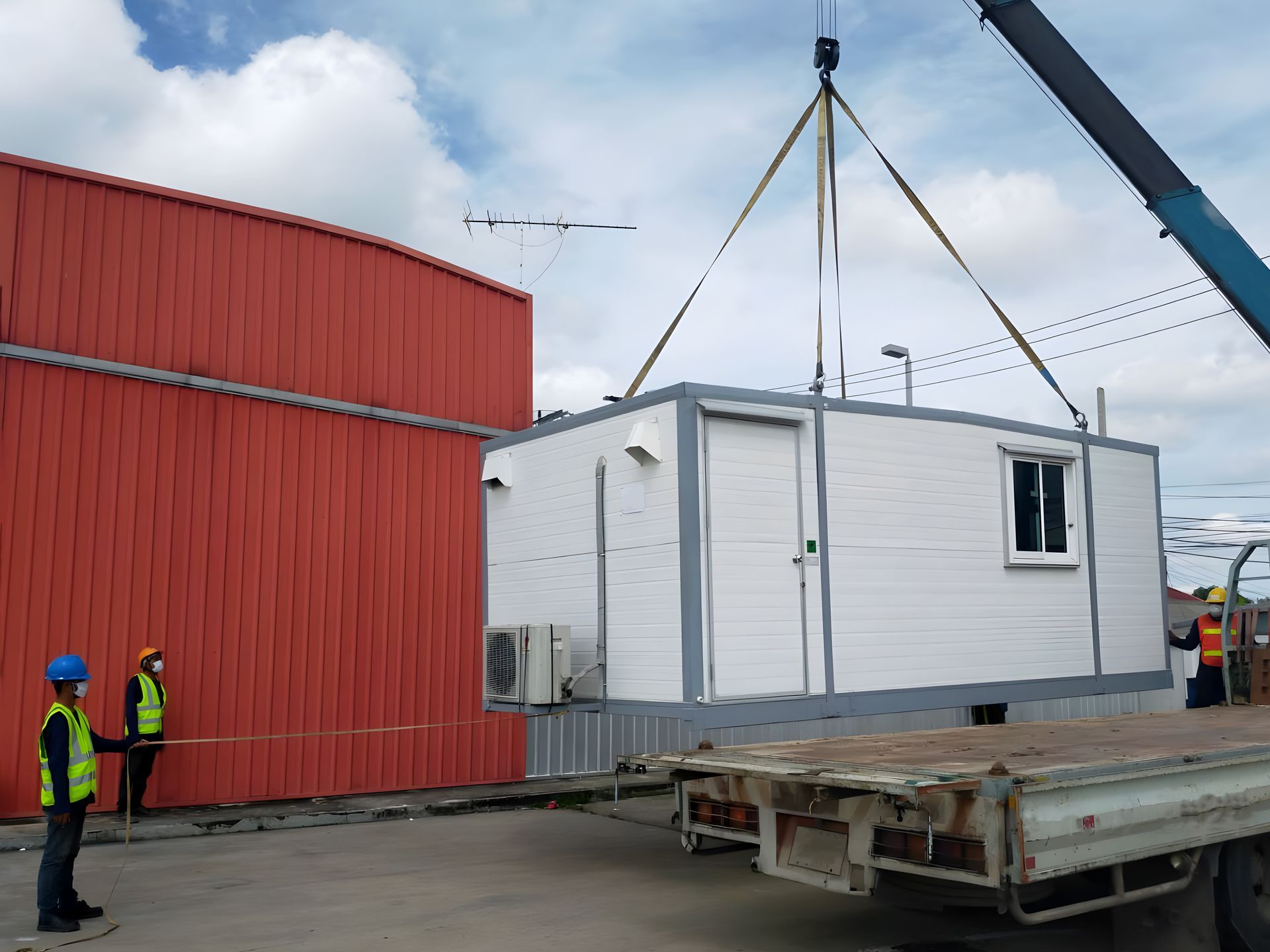 Crane lifting a white modular building near a red industrial structure; two workers watch.