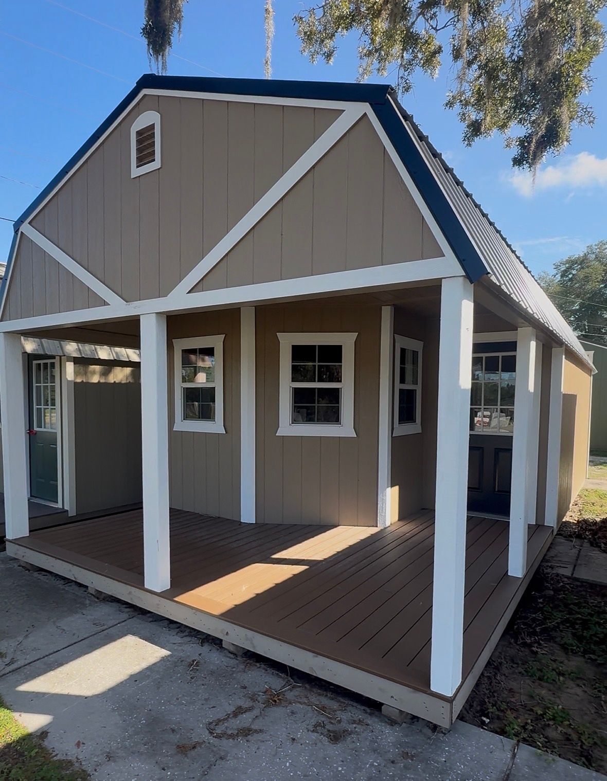 Tan-sided shed with dark roof and white trim, featuring a covered porch with a brown deck and white pillars.
