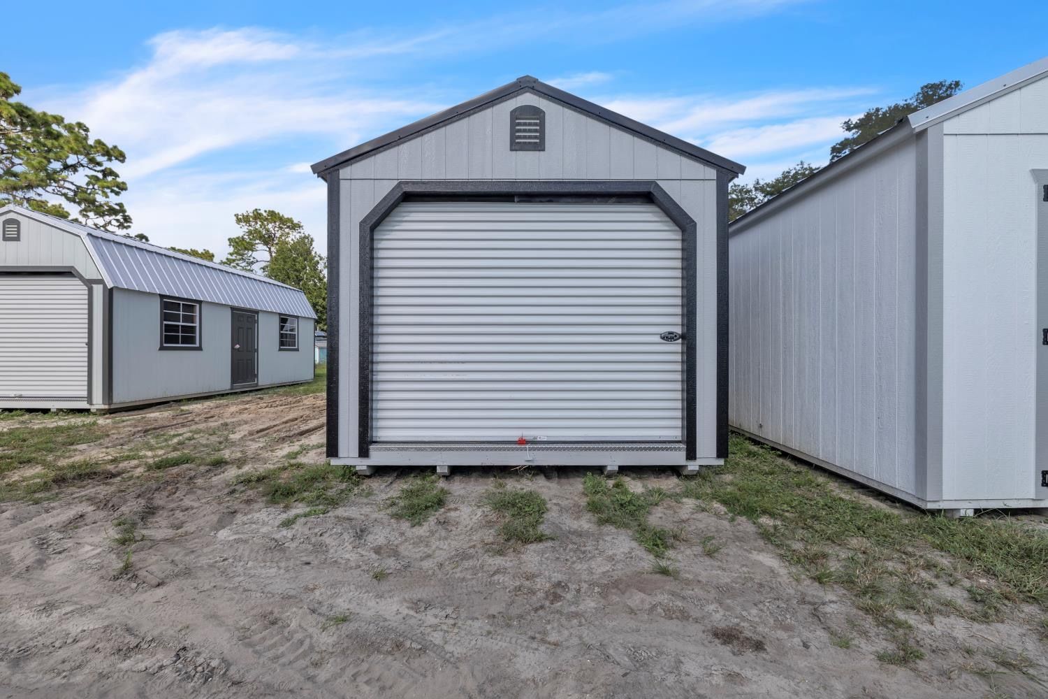 Gray storage shed with roll-up door, black trim, and a peaked roof. Set outdoors on gravel, with other sheds nearby.