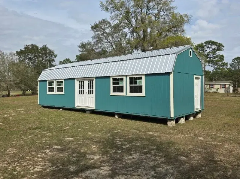 Teal barn-style shed with white trim, metal roof, and windows on a grassy lot.
