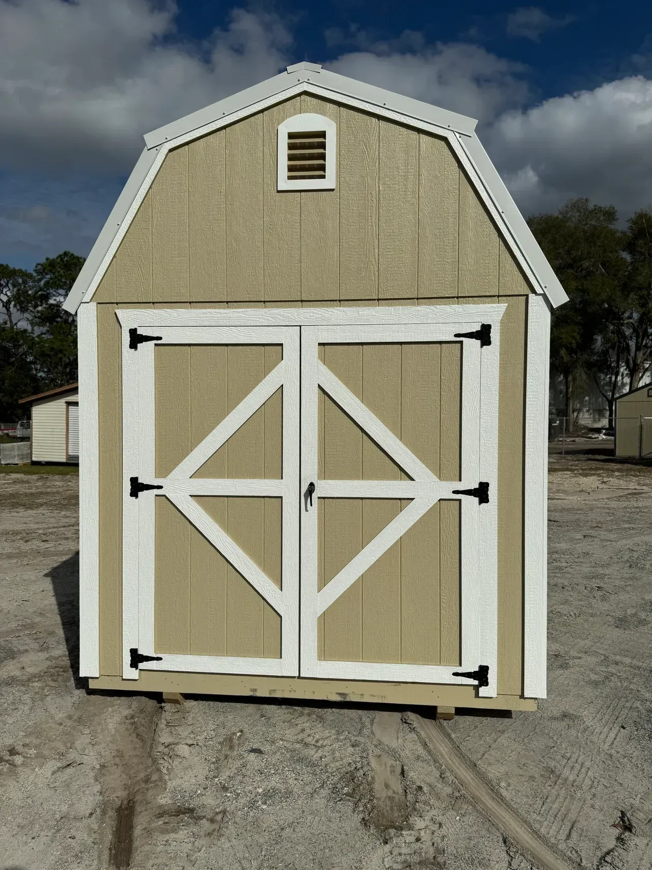 Tan barn-style shed with white trim and X-shaped door accents; small vent above double doors; outdoor setting.