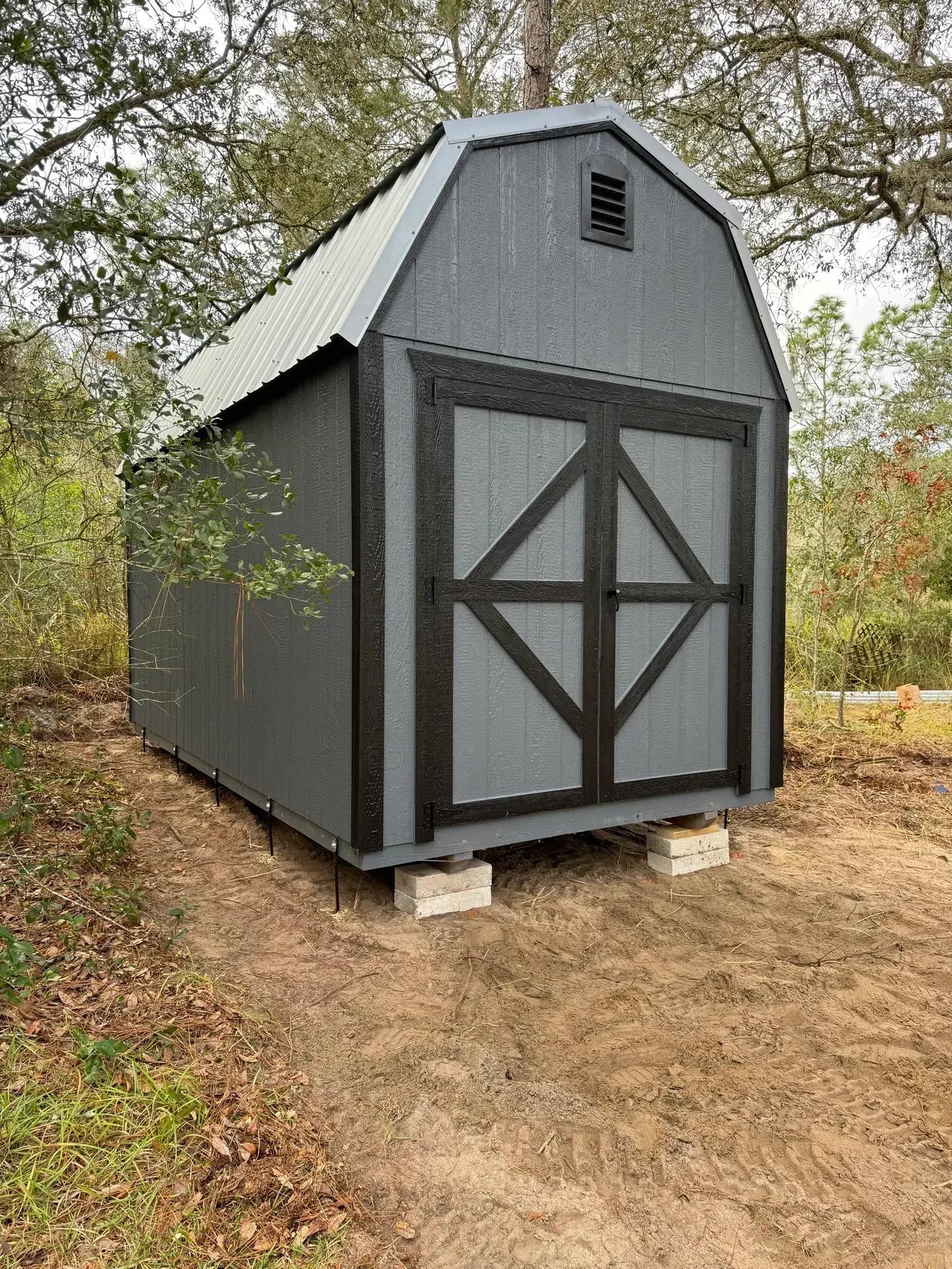 Gray barn-style shed with black trim and doors, silver metal roof, sitting on concrete blocks, outdoors.