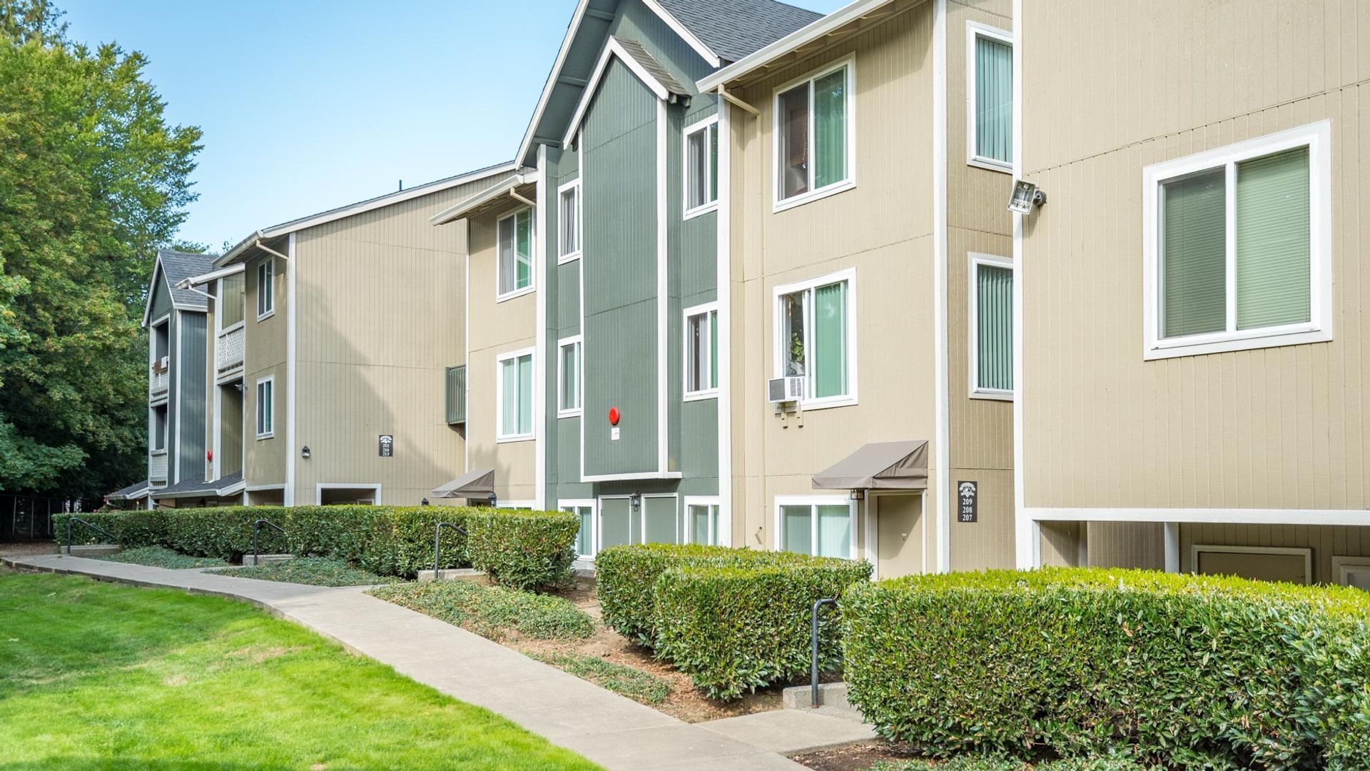 Multi-story apartment building with tan siding, green accents, and manicured green hedges along a paved walkway.