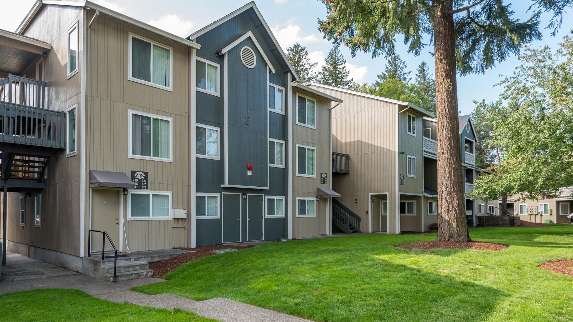 A three-story apartment complex with beige and dark grey vertical siding, surrounded by a lawn and mature trees.