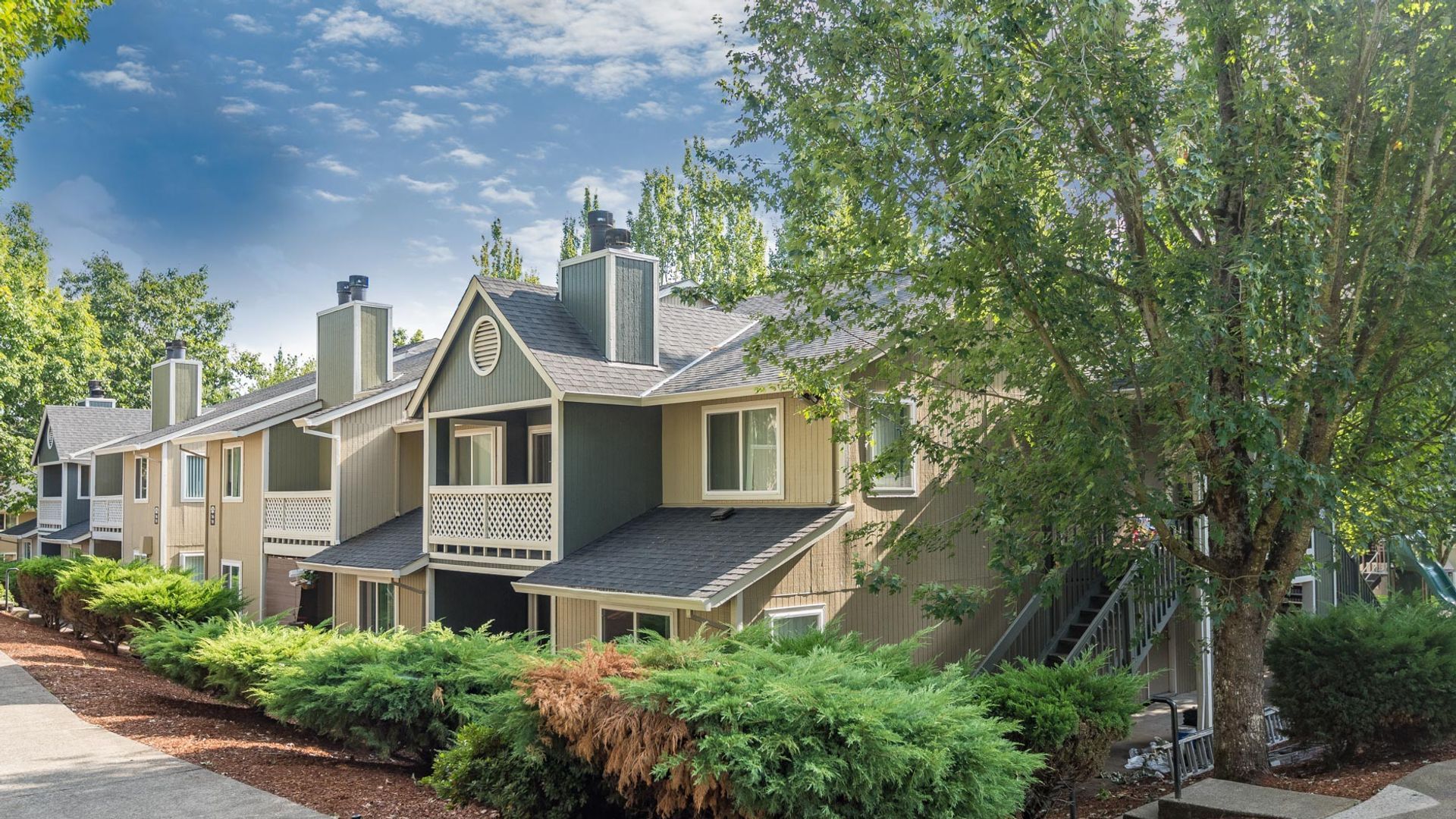 A multi-level apartment complex with beige siding, dark gray trim, and balconies, surrounded by green trees and bushes.