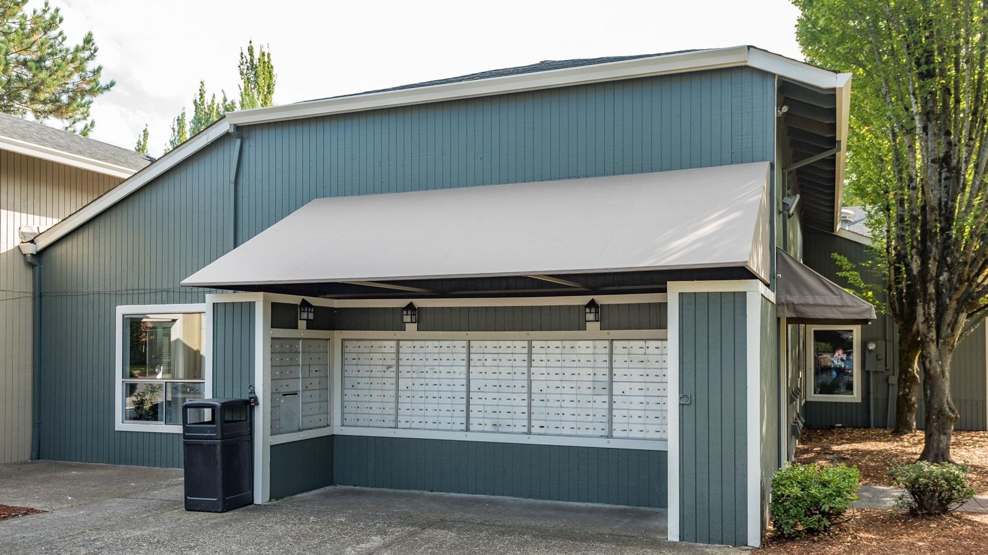 Blue-grey building exterior featuring a central covered kiosk with a large bank of residential mailboxes.