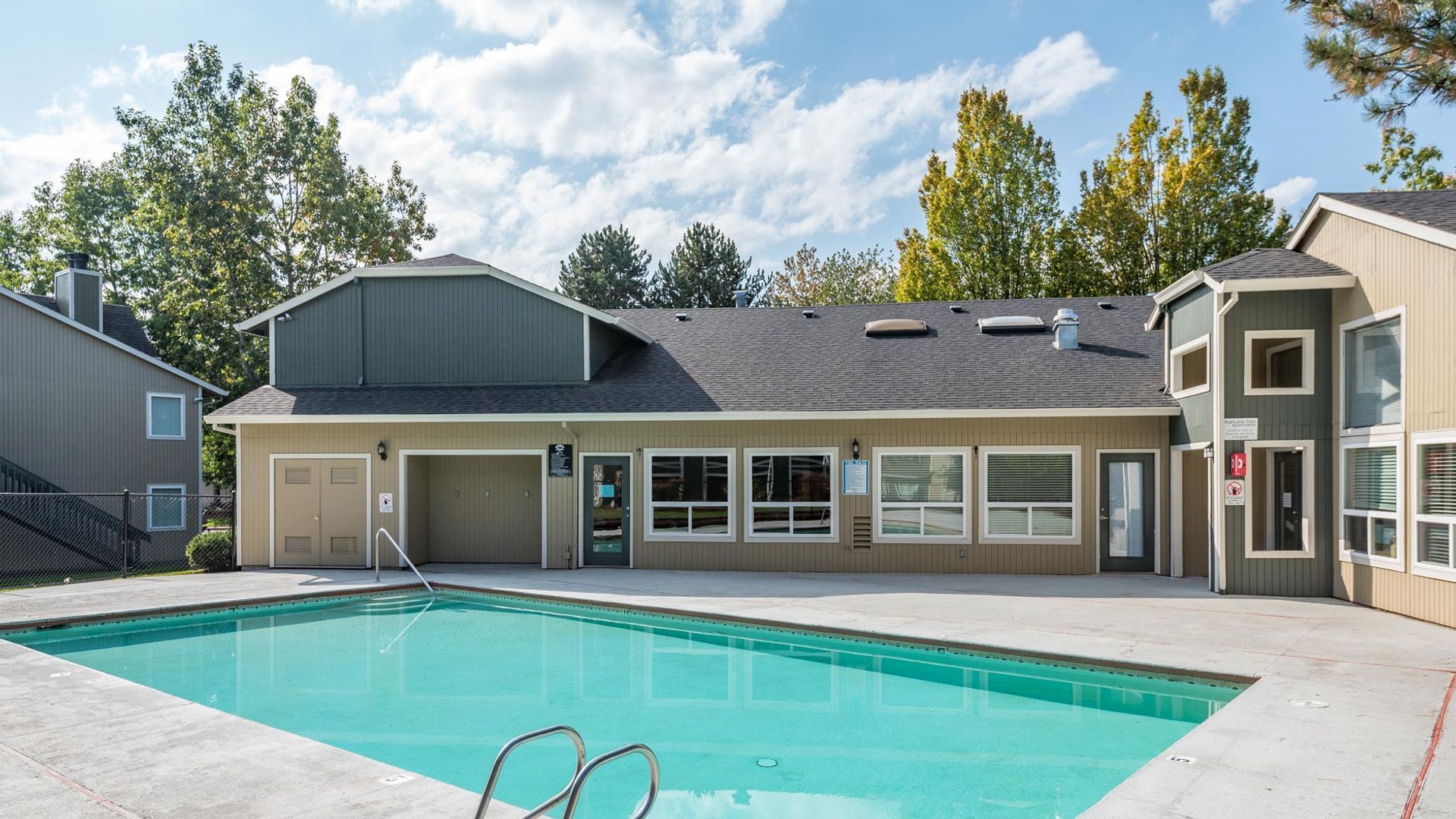 A rectangular swimming pool sits in front of a community building with a shingled roof, trees, and blue sky.
