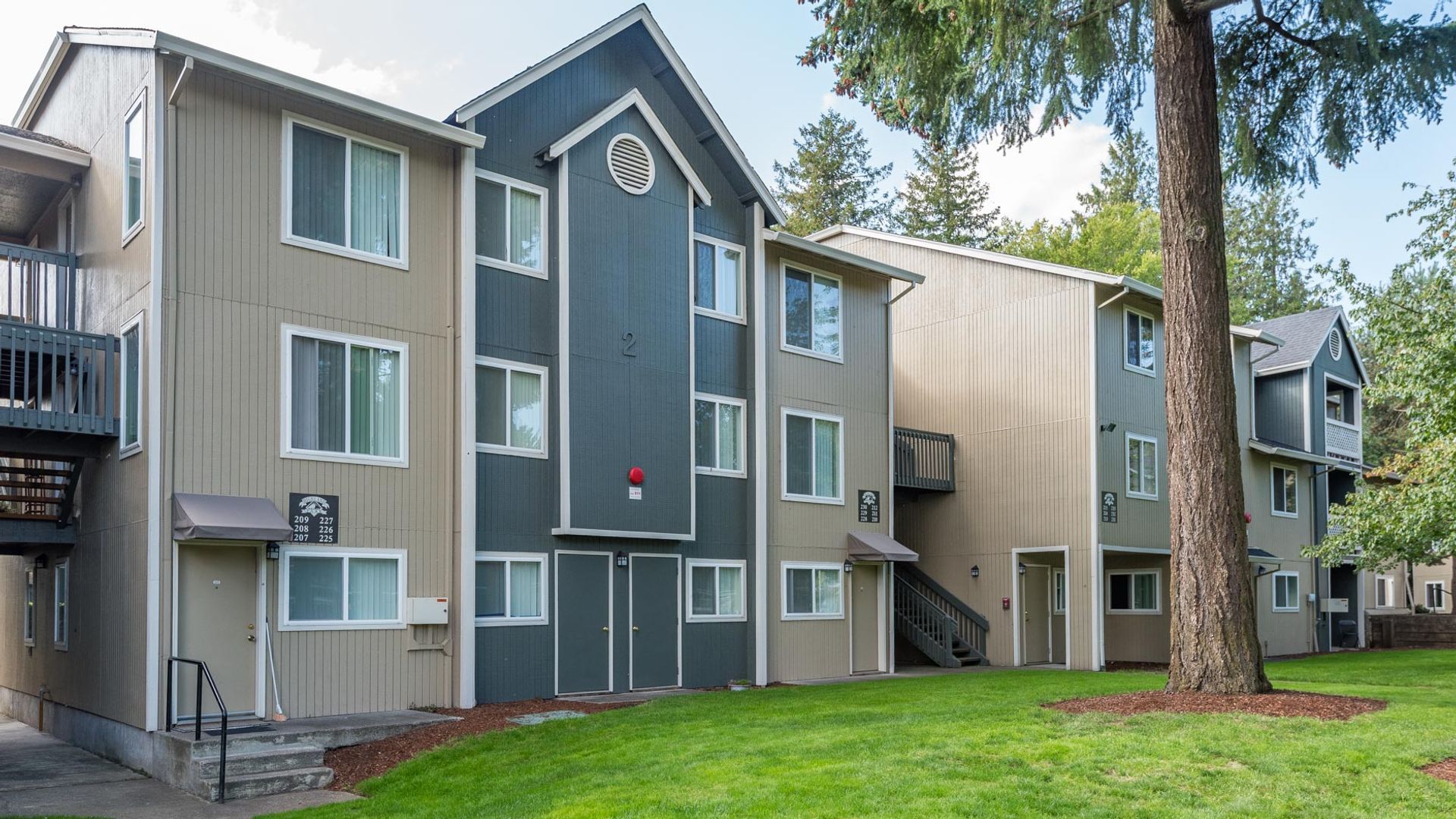A multi-story apartment building with beige and dark blue siding, surrounded by a lawn and mature trees.