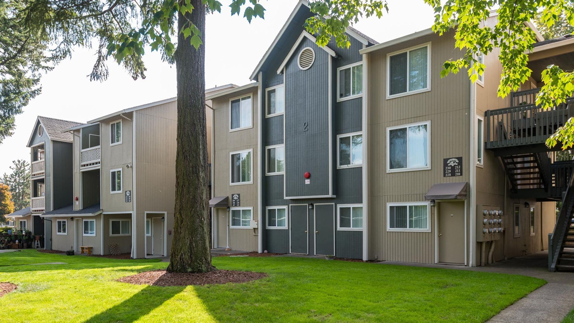 Three-story apartment building with tan wood siding, dark gray accents, and an outdoor staircase, set on a grassy lawn.