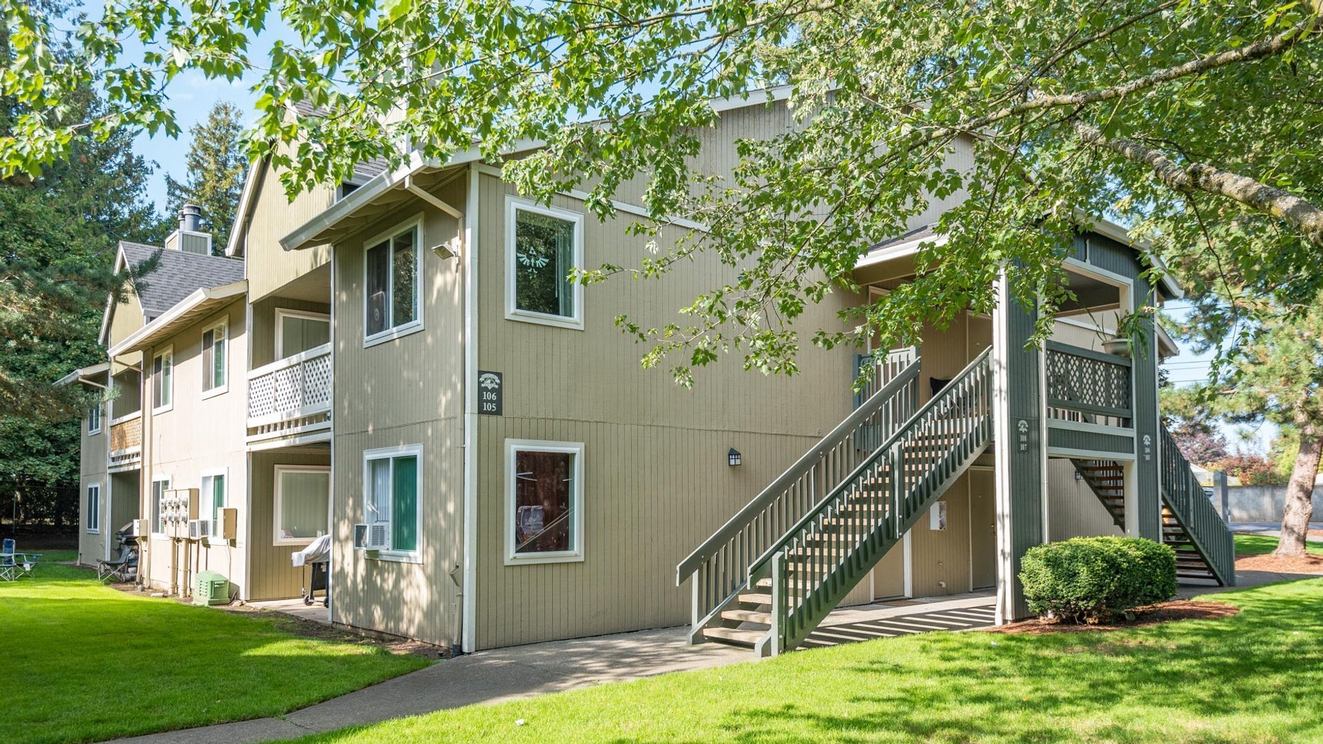 A tan two-story apartment building with wooden stairs, green trim, and surrounding lawn and trees.