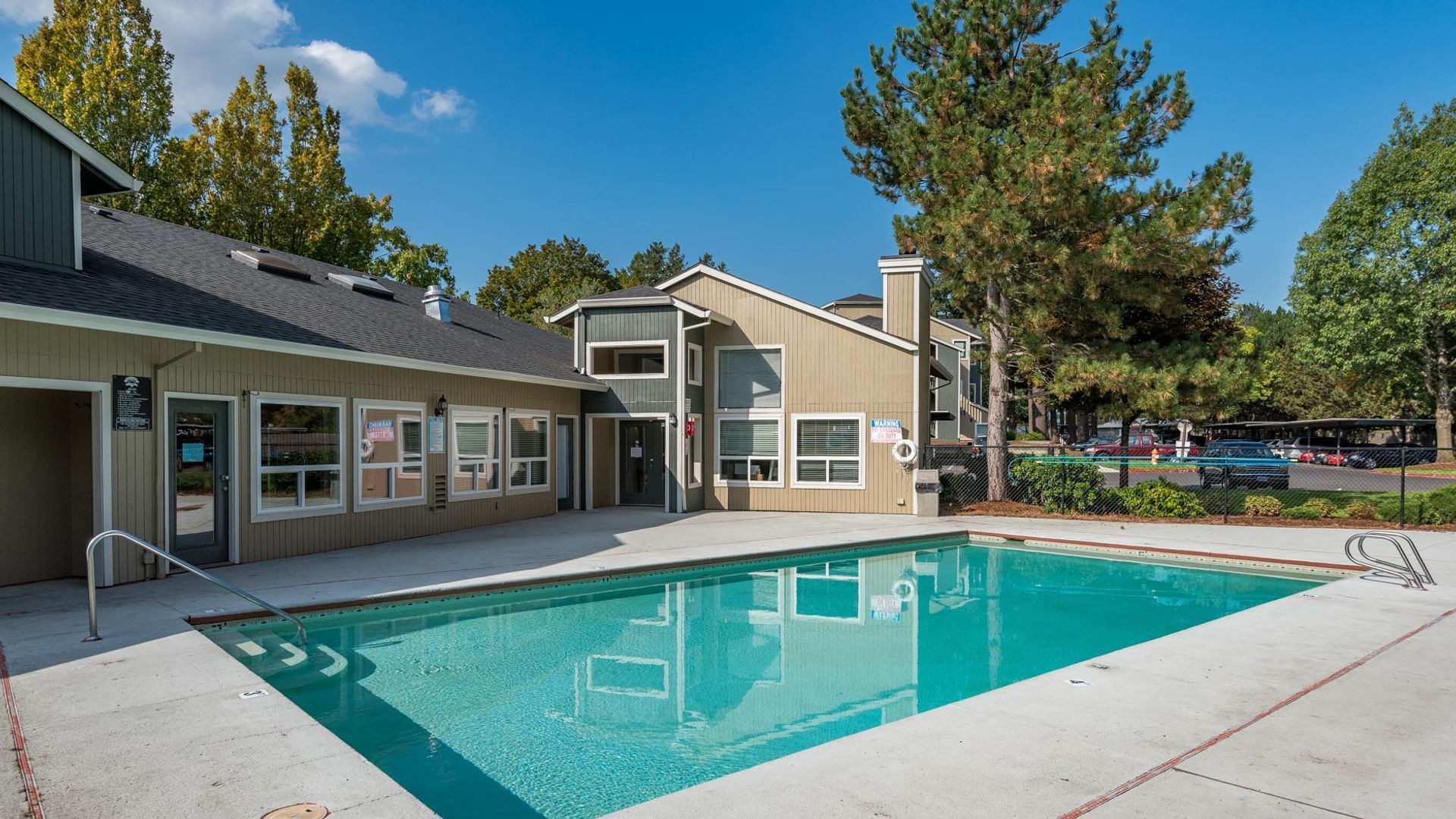 A rectangular outdoor swimming pool with a concrete deck surrounded by apartment buildings and tall trees under a blue sky.