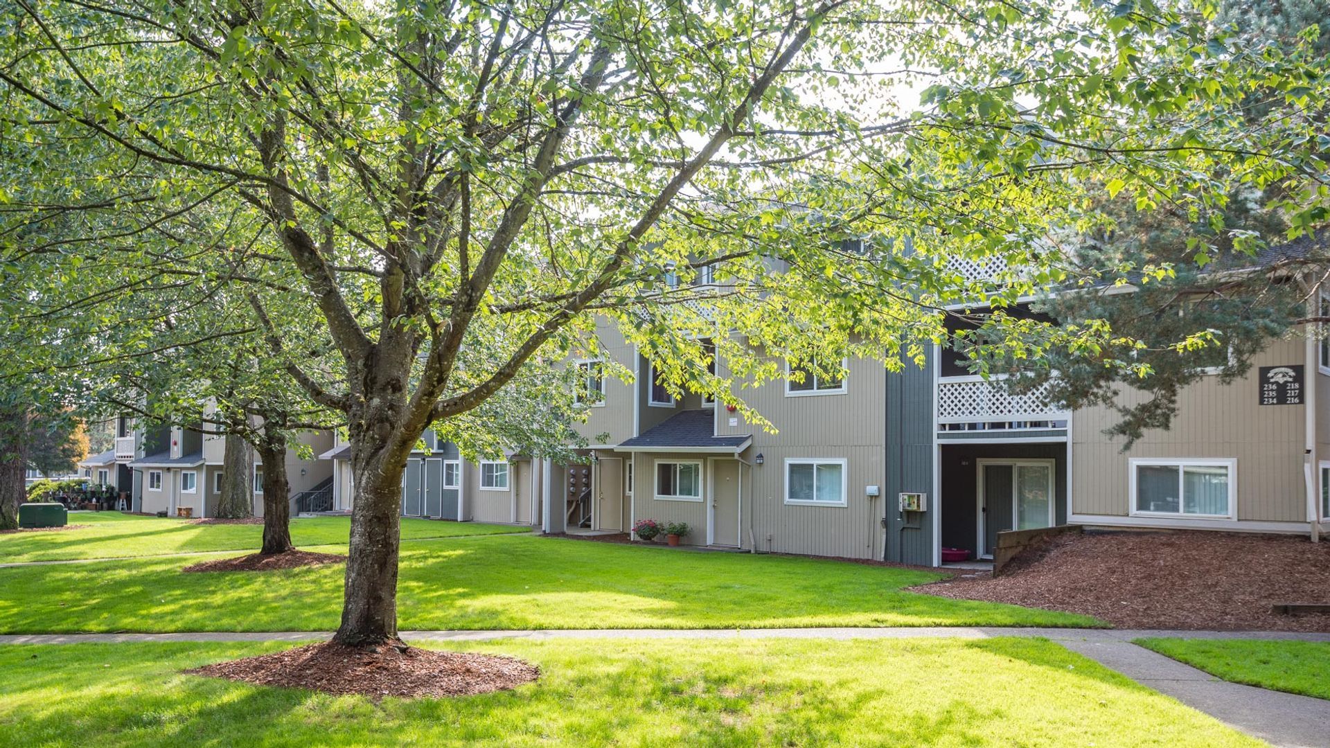 A multi-story apartment complex with beige siding, green lawns, and large trees on a sunny day.