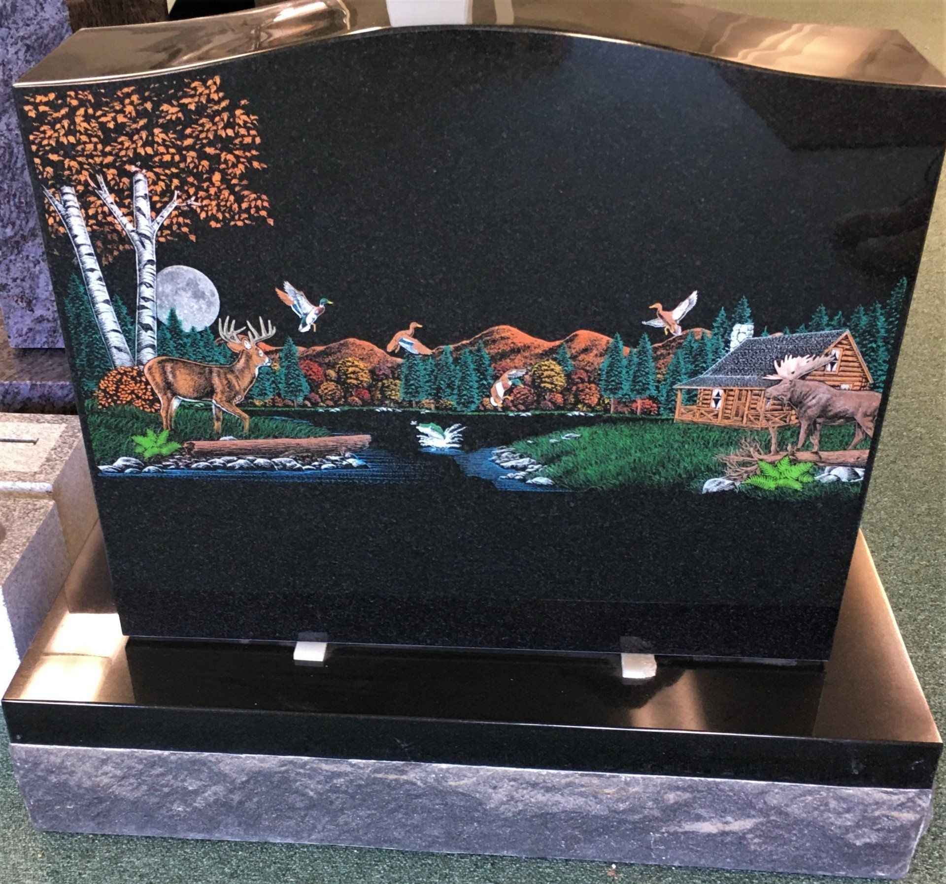 a black gravestone with a colored etching of deer and a cabin on it