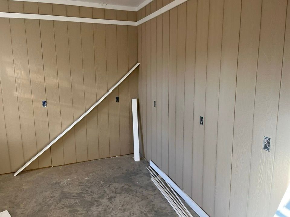 An interior room corner with tan vertical wood-paneled walls, white trim, and scattered baseboard molding pieces.