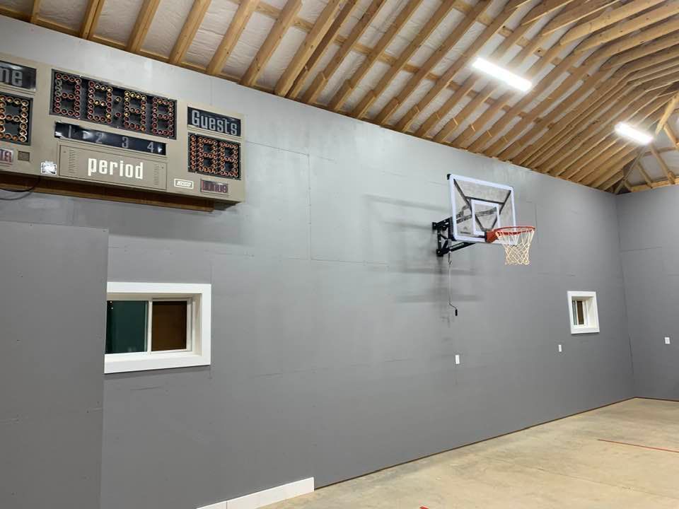 A basketball hoop mounted on a large, grey wall in an indoor court with a digital scoreboard and two windows.