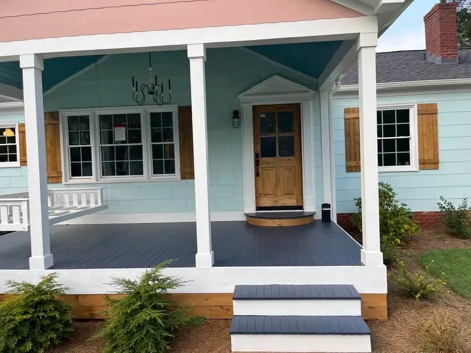 A light blue house exterior features a covered front porch with dark grey decking, white pillars, and a wooden front door.