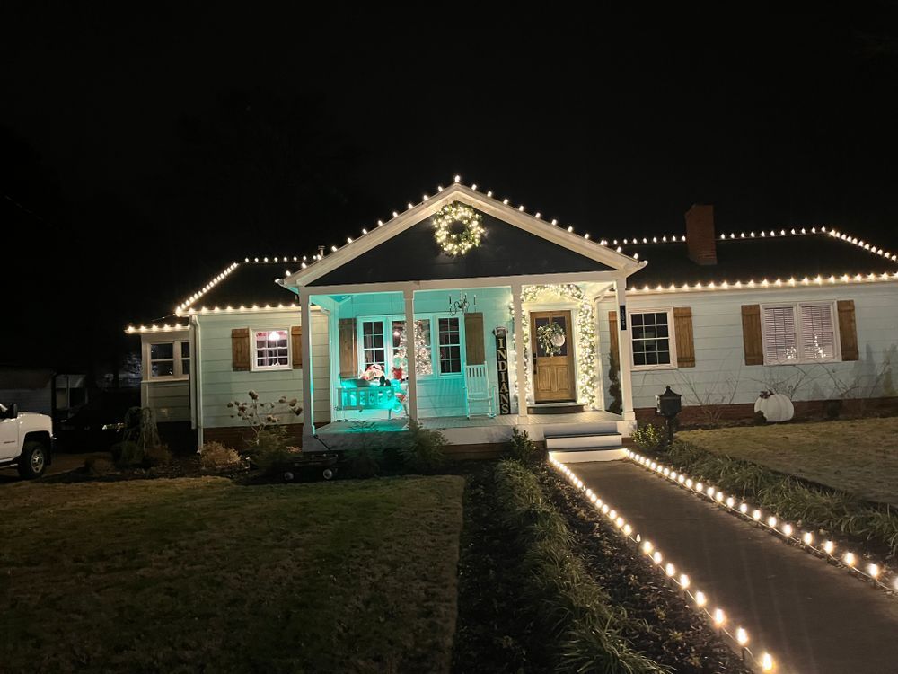 A house at night decorated with string lights along the roof and a path, featuring a lit wreath on the front gable.