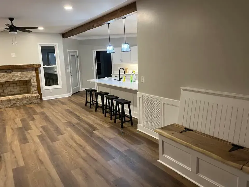 A renovated room with wood-look flooring, a stone fireplace, a kitchen island with bar stools, and a built-in bench seat.