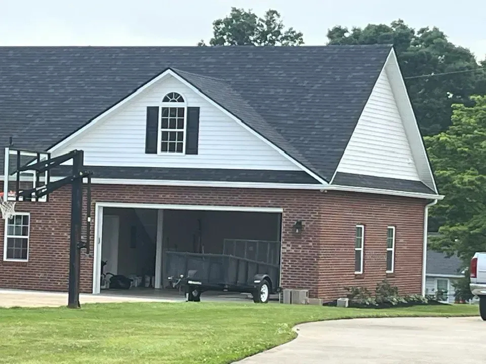 A brick garage with a white upper story and dark roof, with a trailer parked inside and a basketball hoop to the left.