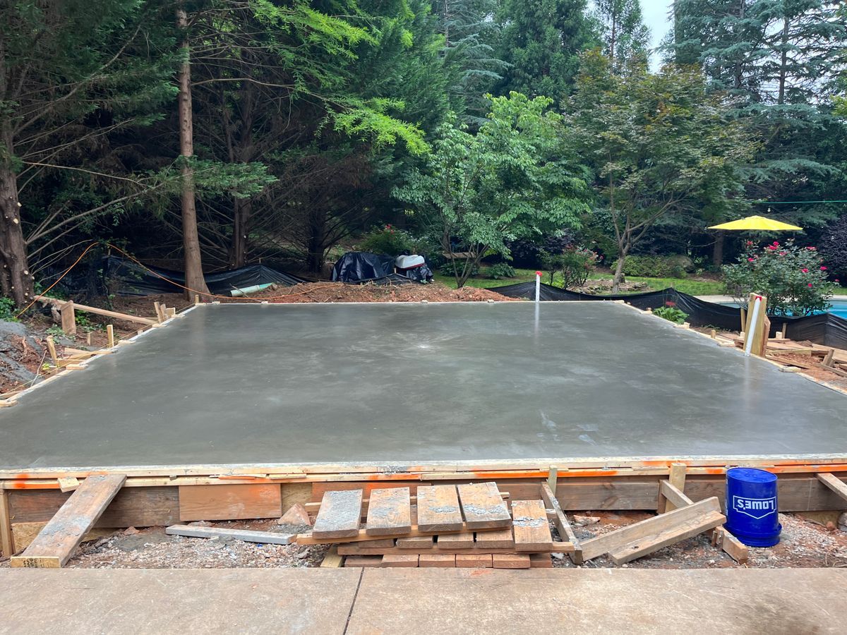 A freshly poured rectangular concrete slab surrounded by wooden forms in a wooded outdoor setting.