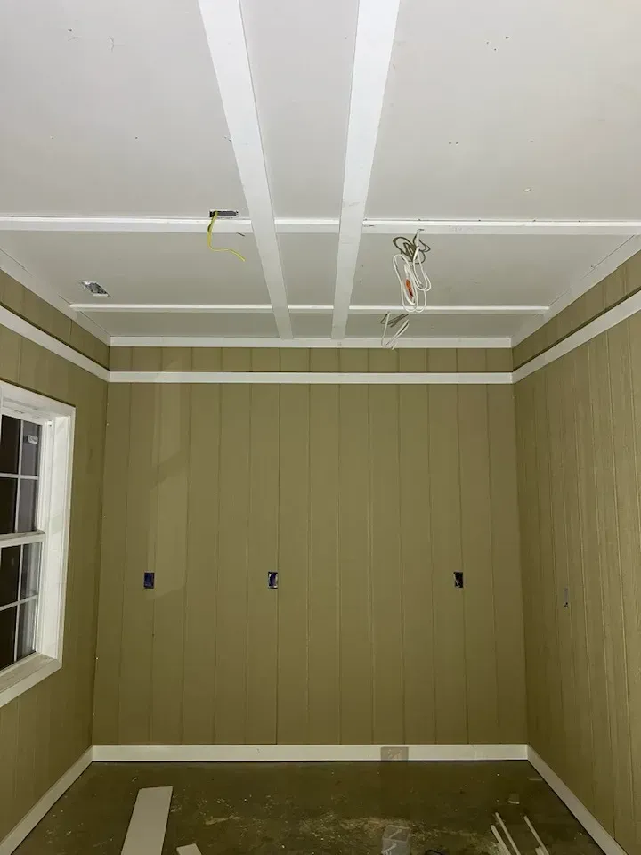 A partially renovated room with tan vertical wood-paneled walls, white trim, and a white grid-patterned ceiling.
