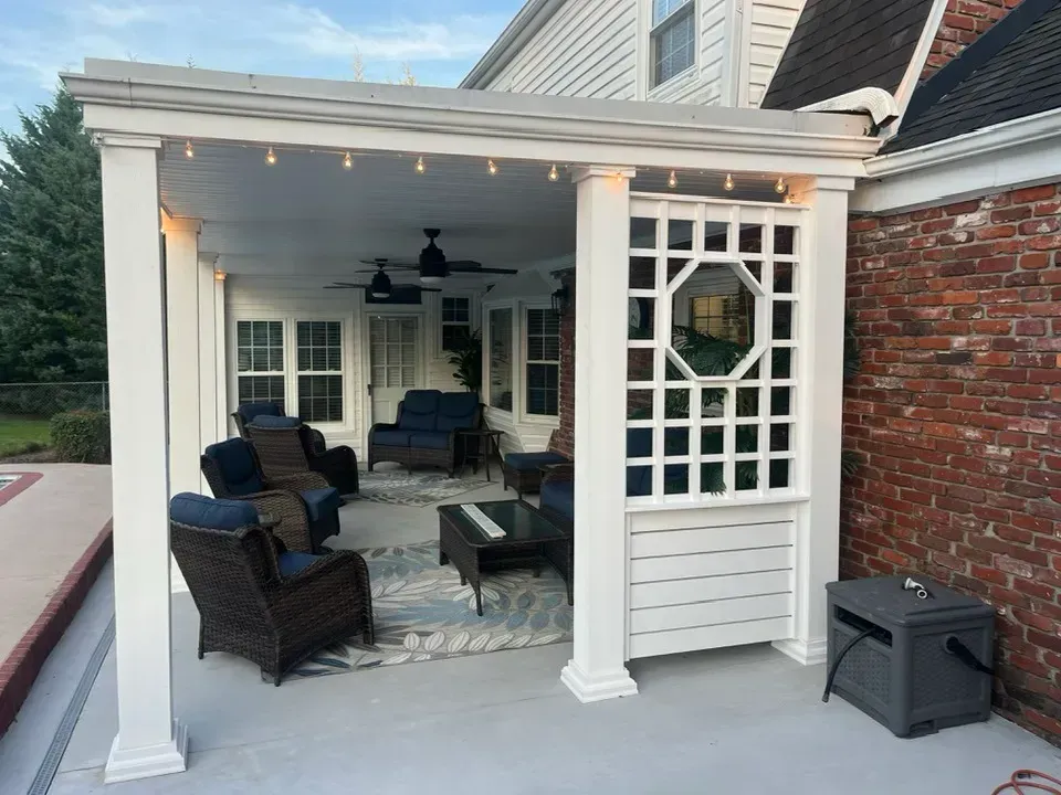 A covered outdoor patio with wicker furniture, a coffee table, and a decorative white lattice wall against a brick house.