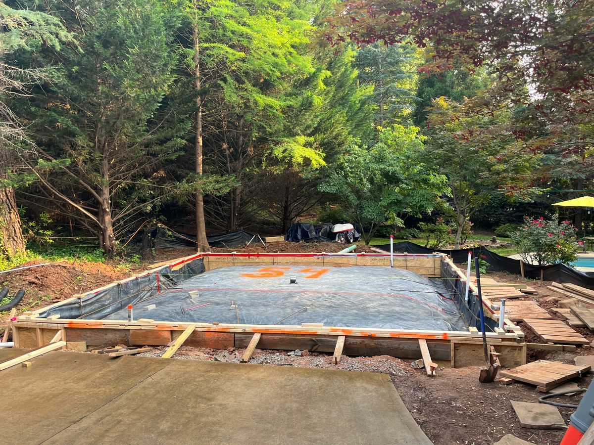 A rectangular excavation site framed with wooden forms, prepared with gravel for a concrete foundation in a wooded yard.