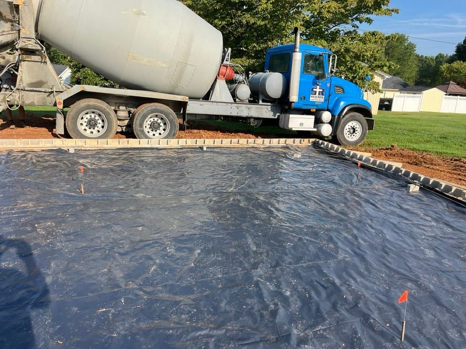 A blue cement mixer truck parked beside a concrete foundation site covered with a plastic vapor barrier.
