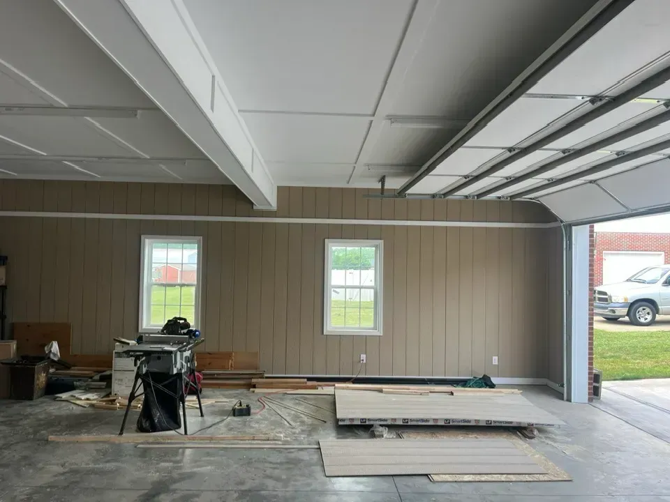 A garage interior under renovation with tan paneling, two windows, a table saw, and wooden boards on the concrete floor.