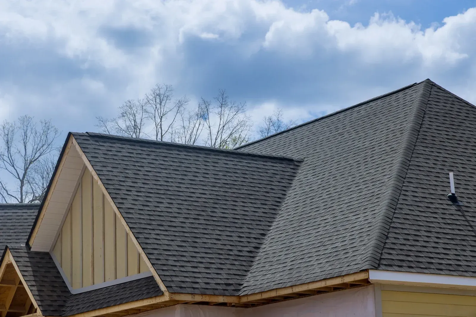The roof of a house under construction with dark grey shingles, light tan siding, and a cloudy blue sky background.