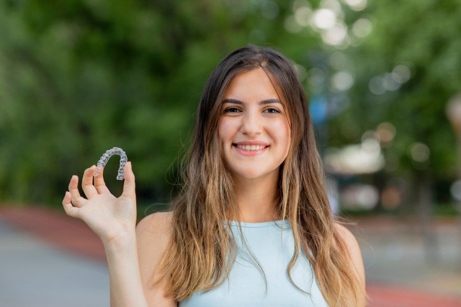 Woman holding clear teeth aligners, smiling. Outdoors, blurred green background.