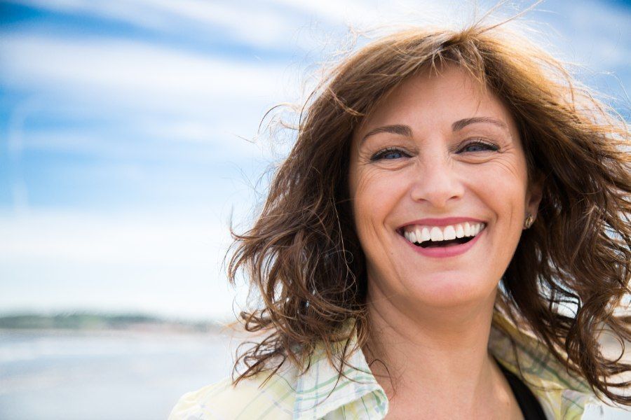 Woman with brown hair smiles broadly outdoors near the ocean.