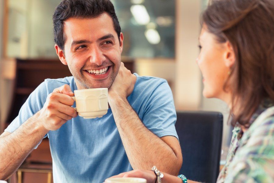 Man smiling and holding a mug, talking to someone; indoors, daytime.
