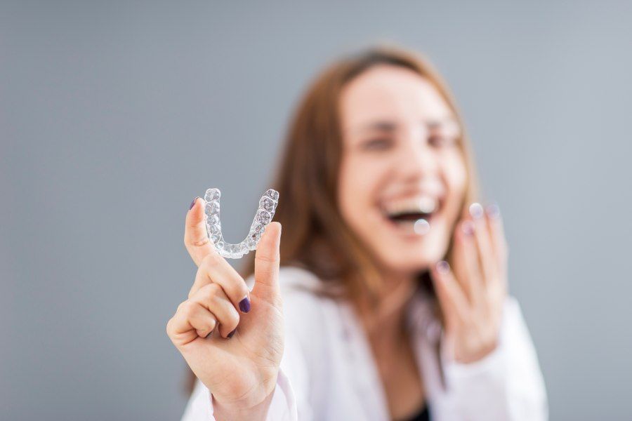 Woman holding clear aligner, smiling, gray background.