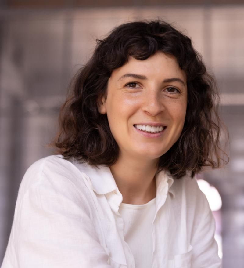 Woman with short, dark curly hair smiles, wearing a white shirt against a neutral background.