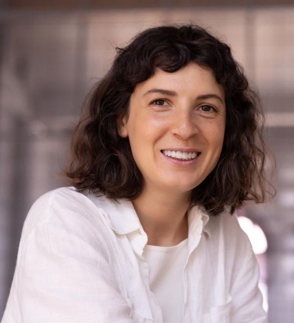 Woman with short, dark curly hair smiles, wearing a white shirt against a neutral background.