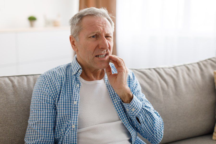 Man seated on couch, grimacing and touching his cheek, possibly due to tooth pain.