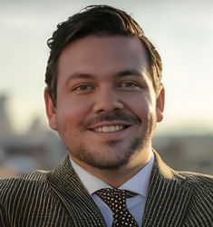 Man in striped blazer and patterned tie smiles outdoors, city background.