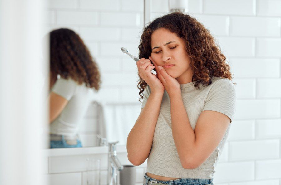 Woman with curly hair holds cheek, grimacing in pain near bathroom mirror. Toothbrush visible.