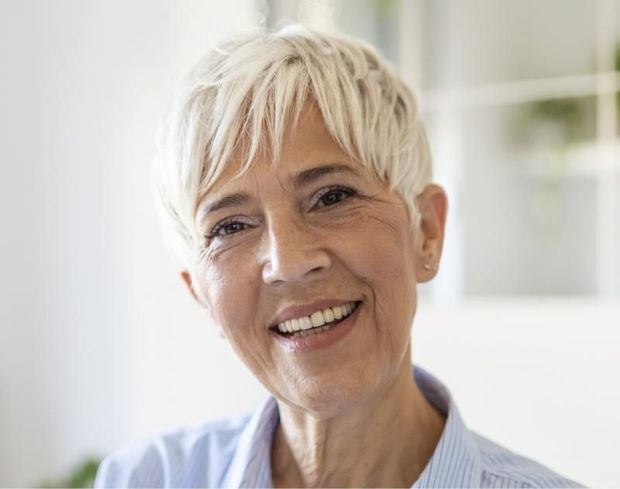 Woman with short, white hair smiles, wearing a blue shirt, indoors.