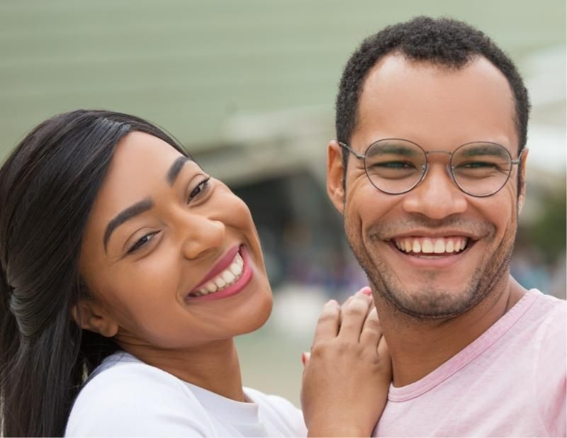 Smiling couple, woman leans on man, both looking at the camera, light background.