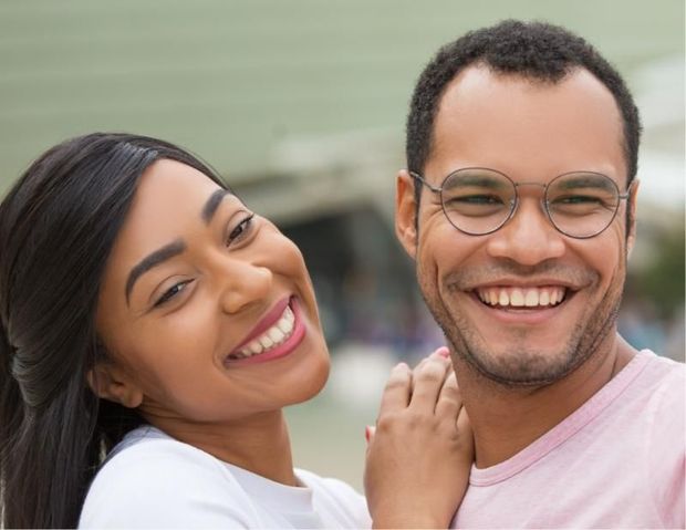 Smiling couple, woman leans on man, both looking at the camera, light background.