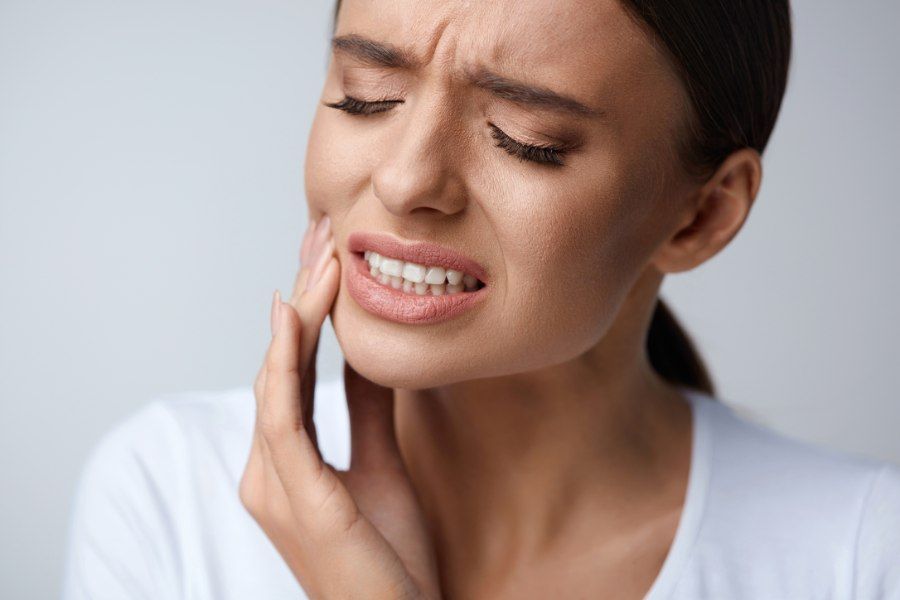 Woman in a white shirt, grimacing, holding her cheek in apparent pain.
