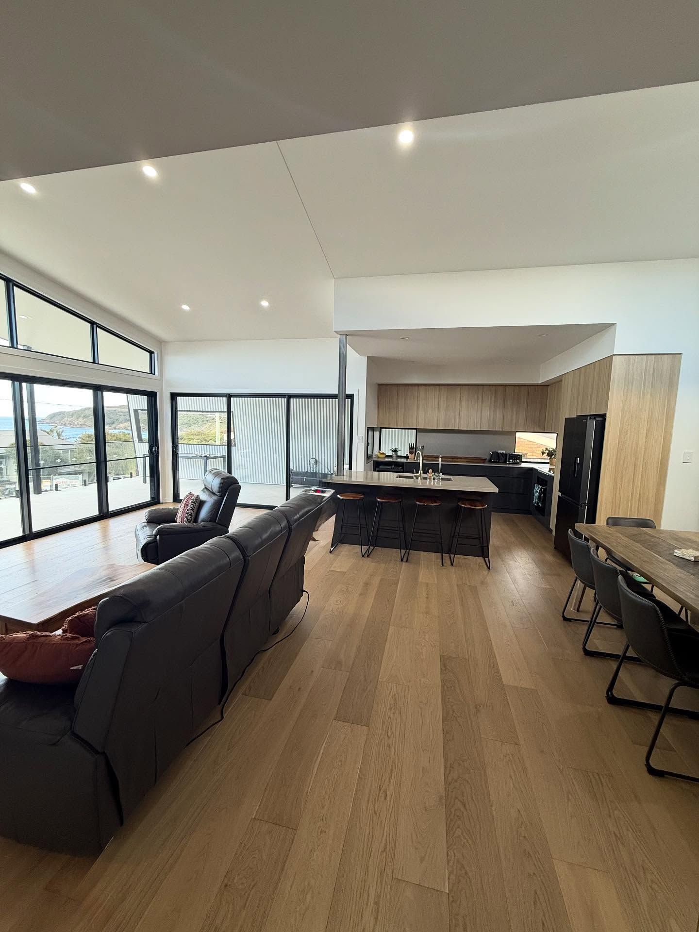 Open-concept living space with kitchen island, dark furniture, wooden floors, large windows, and bright lighting. — Barrington Coast Homes in Old Bar, NSW