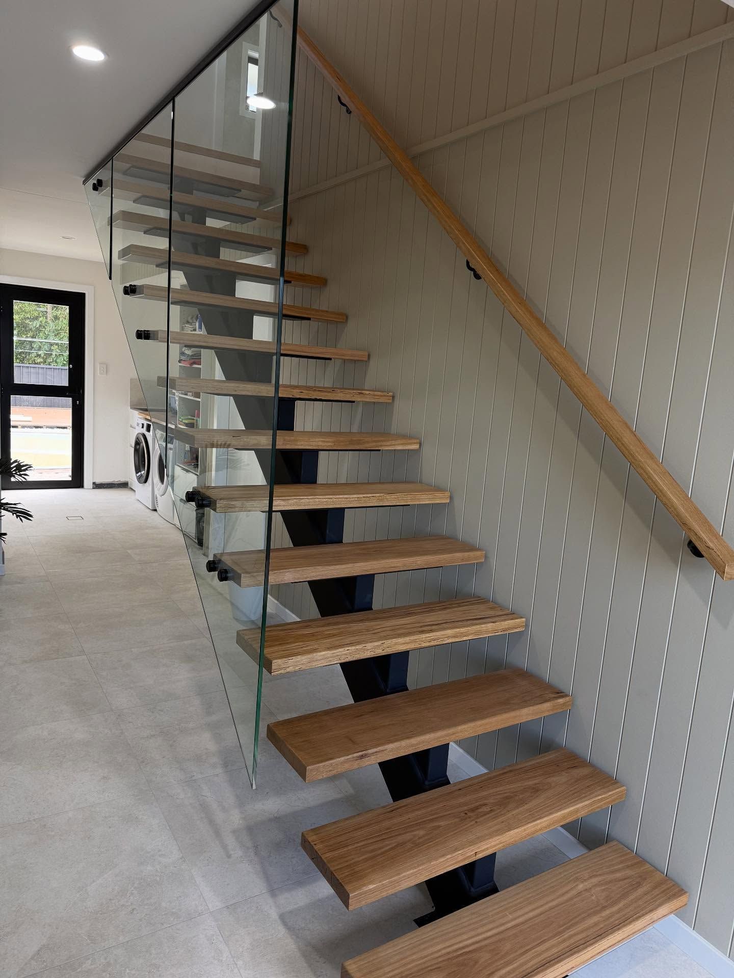 Wooden floating staircase with glass railing and wooden handrail against a light grey wall  — Barrington Coast Homes in Tallwoods, NSW