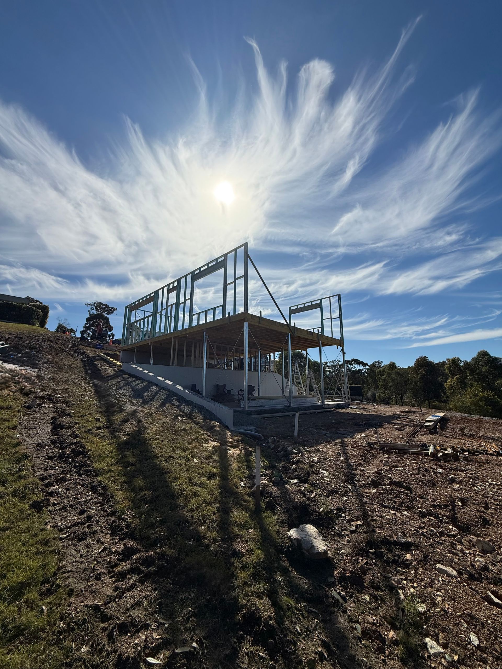 Steel frame of a house under construction on a hillside, bright blue sky with wispy clouds, sunny. — Barrington Coast Homes in Tallwoods, NSW