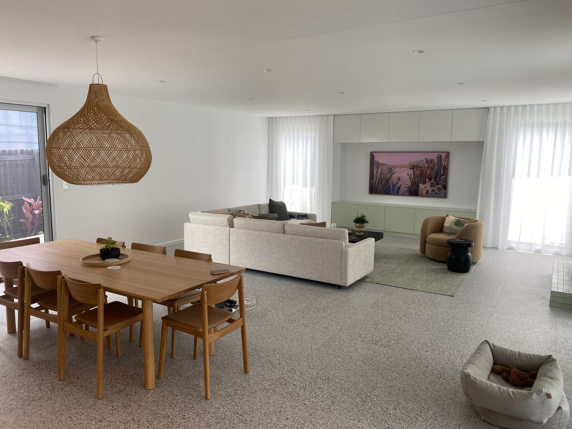 Open-plan living room with dining and seating areas. Beige sofa, wooden table and chairs, tan accent chair, white walls, and terrazzo floors. — Barrington Coast Homes in Forster, NSW