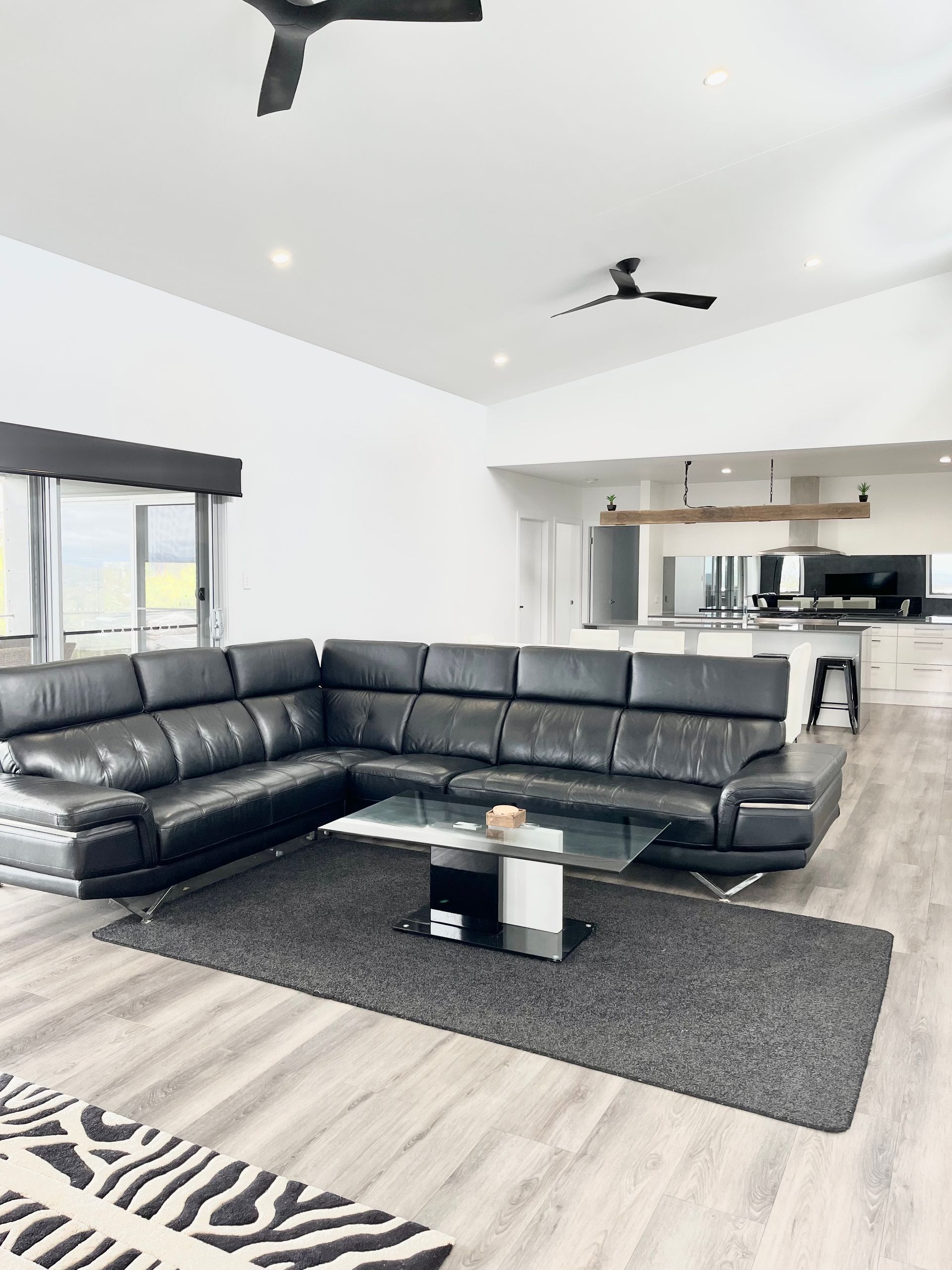 Modern living room with black sectional sofa, rug, and glass coffee table. White walls, wood floor, open kitchen  — Barrington Coast Homes in Tallwoods, NSW