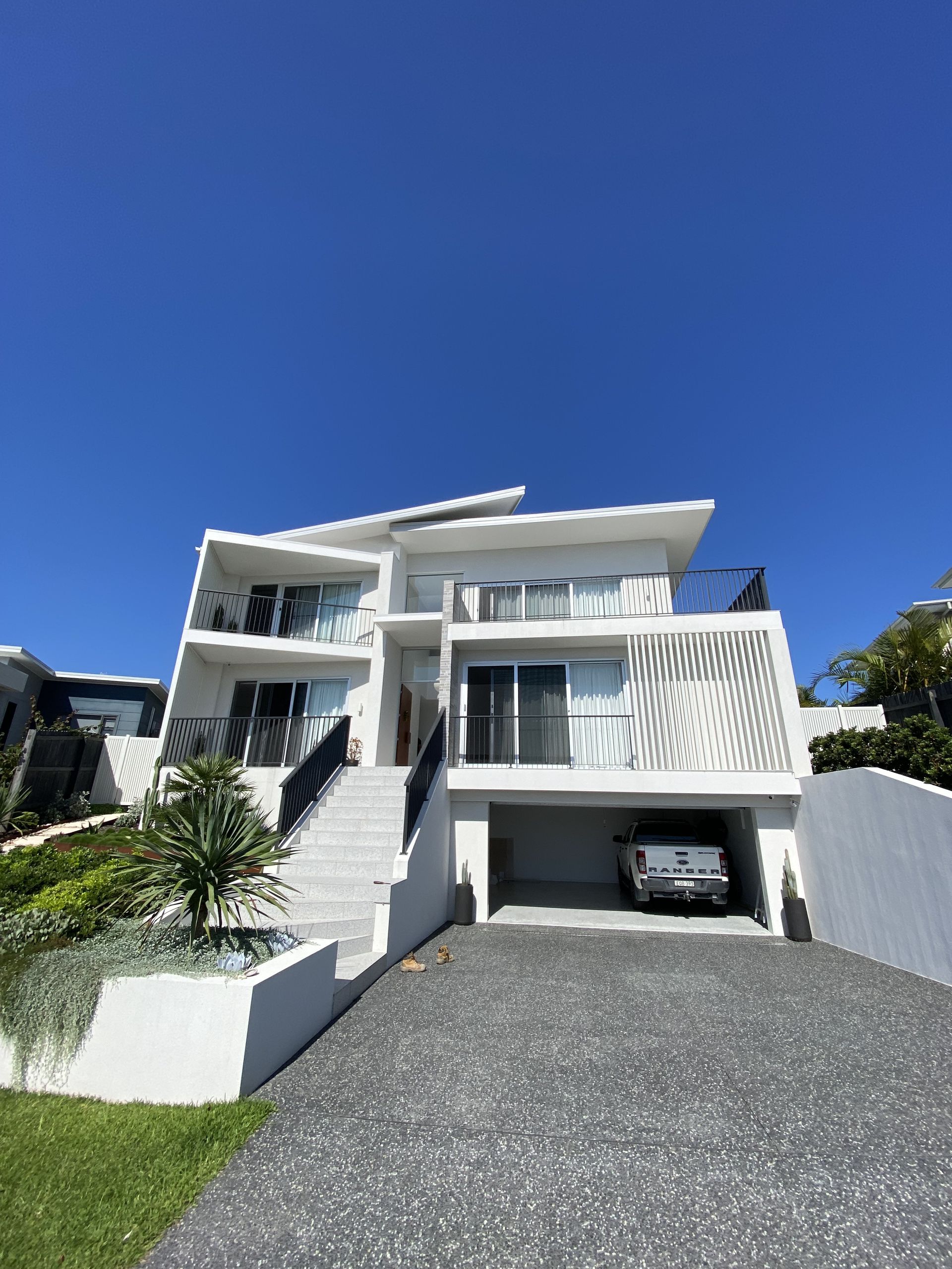 White modern house with a garage, stairs, and a car in the driveway on a sunny day. — Barrington Coast Homes in Tallwoods, NSW